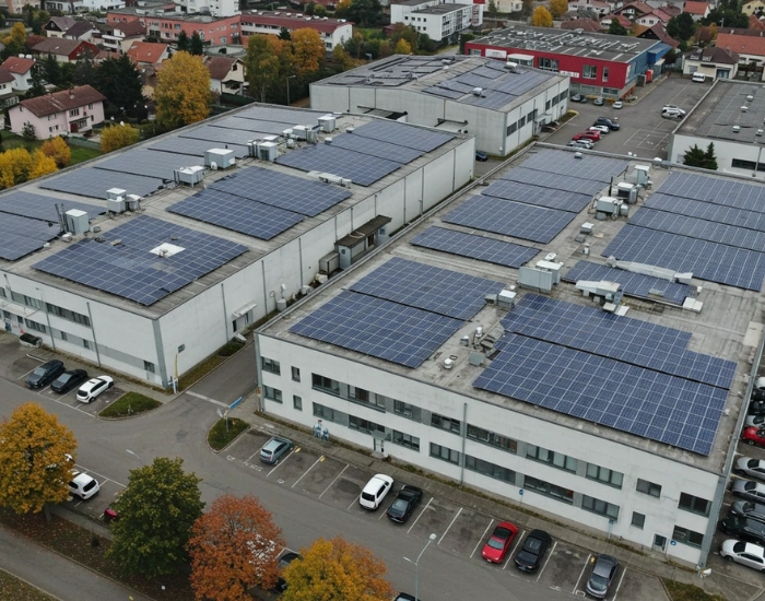 Aerial view of an industrial complex with white buildings featuring rooftop solar panels and a parking lot with vehicles.